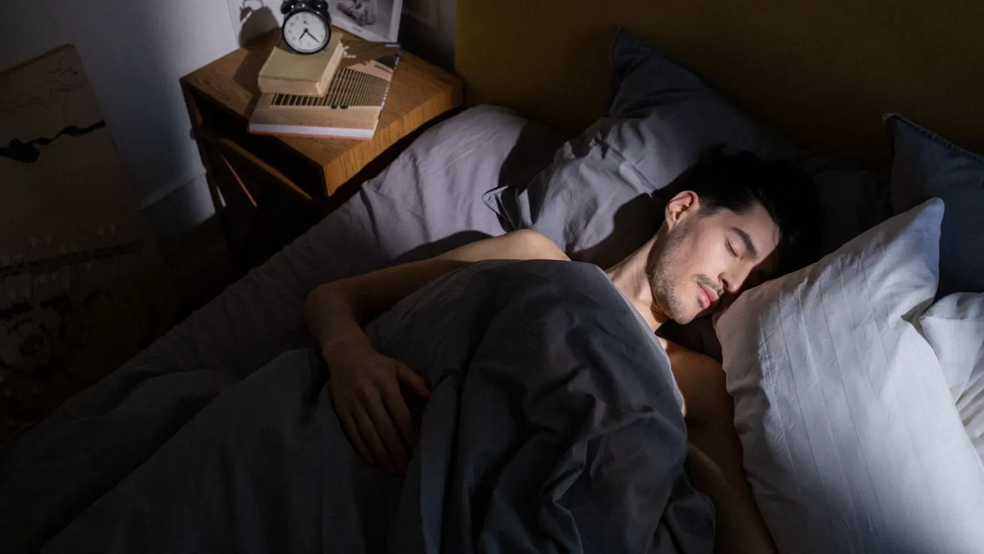 Hombre dormido de lado en una cama con sábanas grises, con un reloj despertador y libros sobre la mesa de noche: imagen para la guía de cómo mejorar la erección, que empieza por el sueño