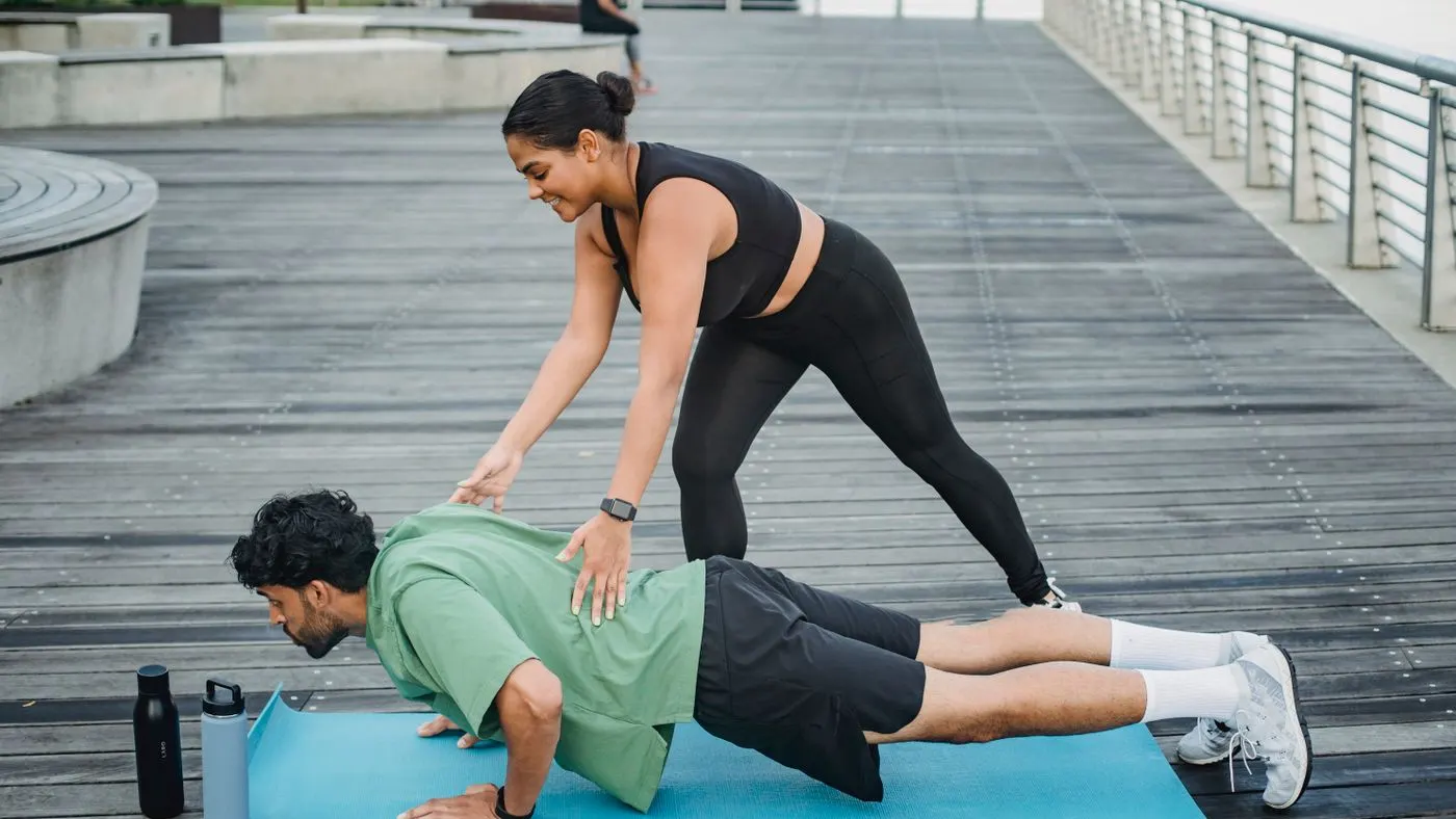 Hombre haciendo flexiones sobre una colchoneta azul en un muelle de madera, con una mujer que lo guía desde atrás y dos botellas de agua al lado: imagen para la guía de cómo durar más en la cama con técnicas y entrenamiento