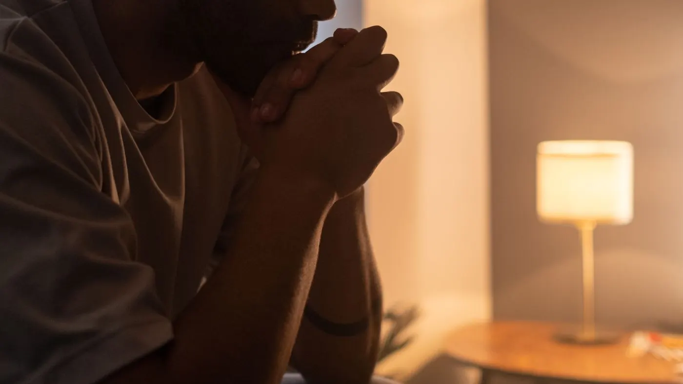 Hombre sentado de perfil en una habitación en penumbra, con las manos juntas frente a la boca y una lámpara encendida al fondo: imagen para la guía sobre la falta de deseo sexual en el hombre