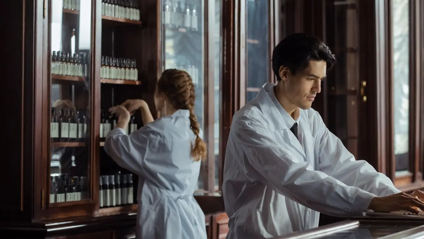 Dos farmacéuticos con bata blanca trabajan tras el mostrador de madera de una droguería antigua, con frascos en las vitrinas: imagen para la guía del sildenafil
