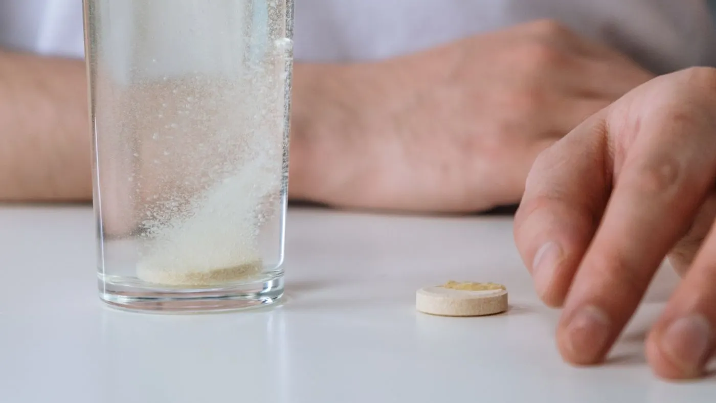 Tableta junto a un vaso de agua con otra tableta disolviéndose sobre una mesa blanca, con las manos de un hombre al fondo: imagen para la guía del tadalafilo