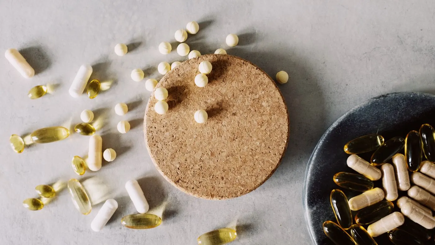Cápsulas blancas, perlas amarillas y cápsulas beige de suplementos regadas sobre una mesa gris junto a un posavasos de corcho y un plato oscuro: imagen para la guía de vitaminas y minerales para la erección